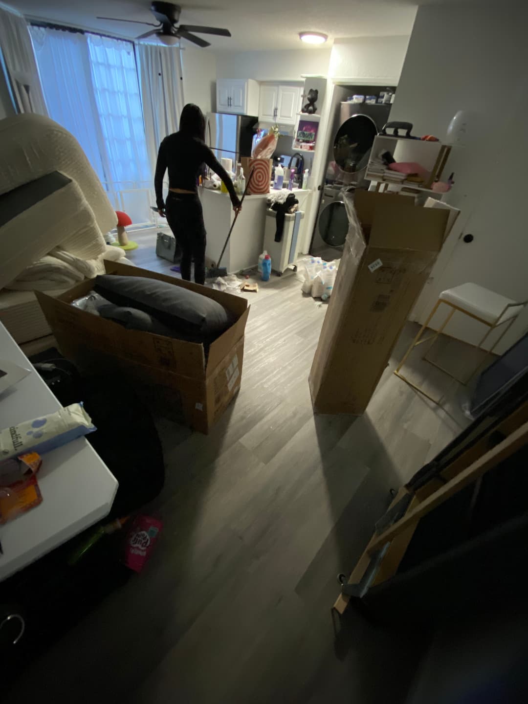 Person cleaning a cluttered apartment with cardboard boxes and furniture scattered around.