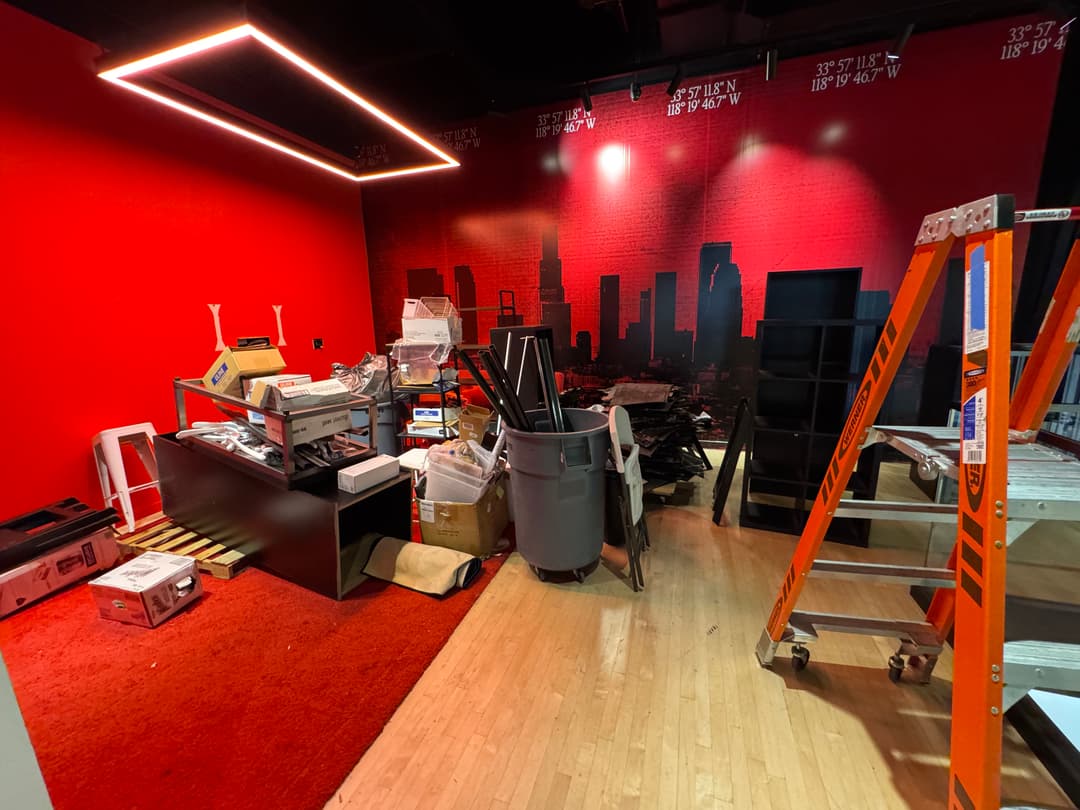 Cluttered workspace with boxes, furniture, and a ladder against a red wall with skyline mural.