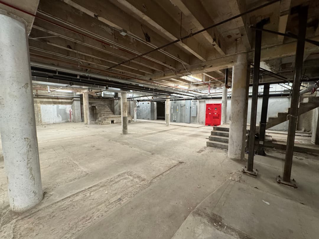 Empty basement interior with exposed beams, concrete floor, and red fire exit doors.