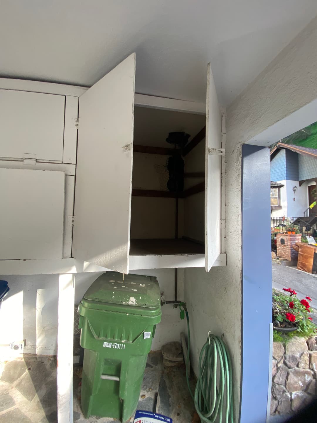 Empty white storage cabinets in a garage with a green trash can and garden hose visible.