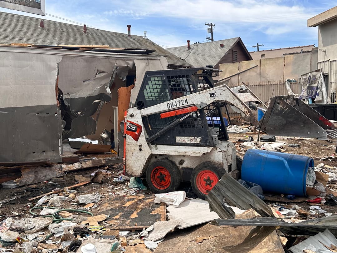 Bobcat skid steer parked among debris and wreckage at a damaged property site.