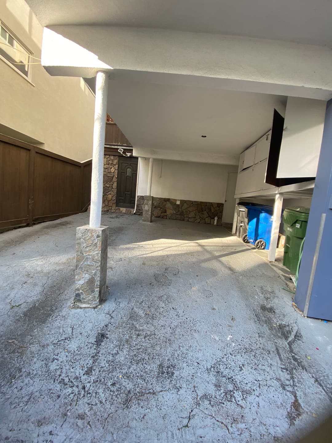 Spacious carport area with stone pillars, garage door, and green recycling bins.