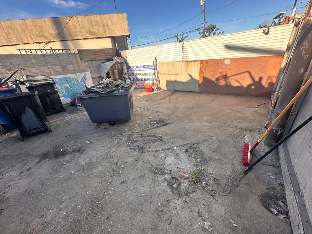 Vacant urban lot with trash bins, debris, and a rake, under clear blue sky.