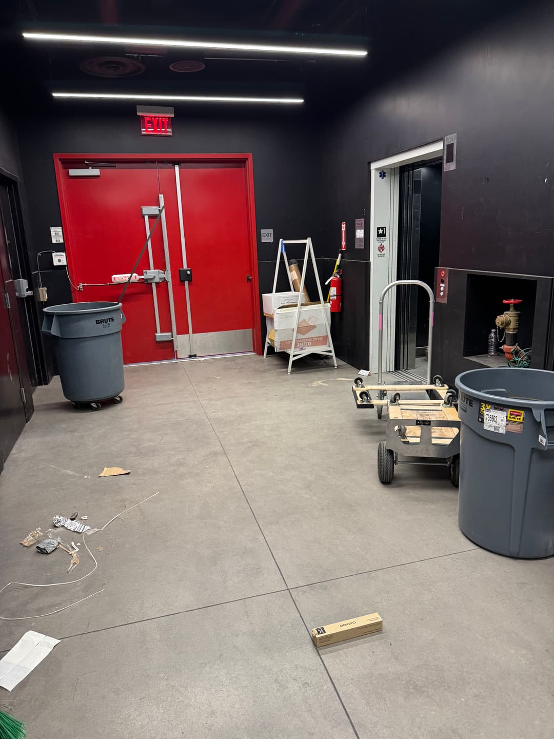 Interior hallway with red door, trash bins, and a cart near an elevator. Construction items scattered.