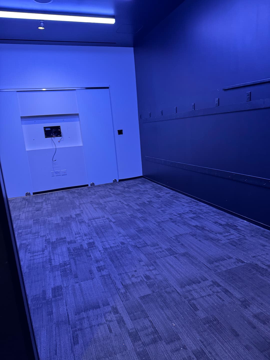 Empty blue-lit room with gray carpeting and a wall-mounted monitor.