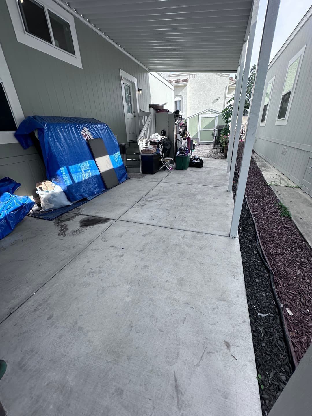 Sidewalk view of a covered patio area with storage items and landscaping details.