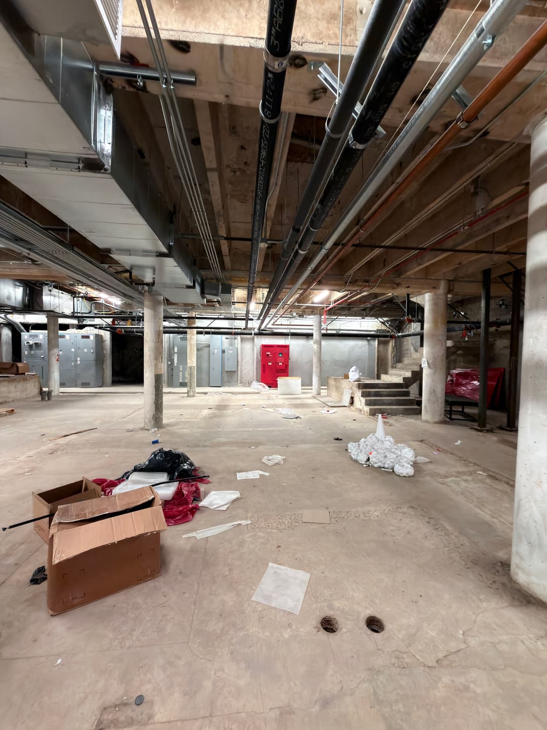Construction site basement with exposed pipes, debris, and a red emergency door.