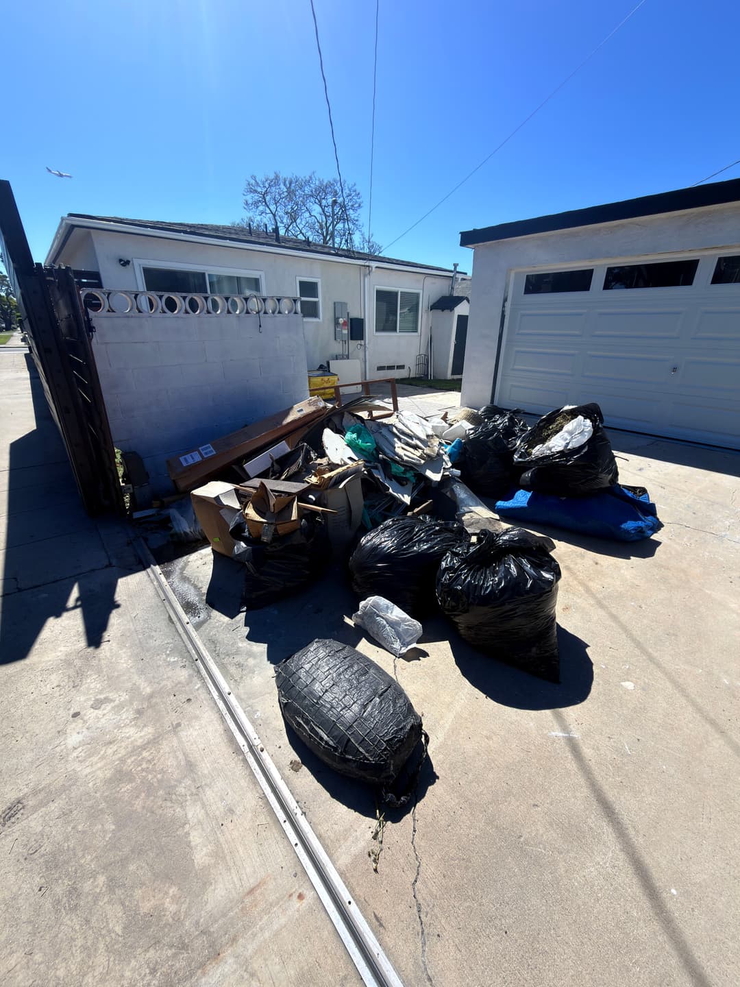 Piles of black garbage bags and discarded items beside a garage on a sunny day.