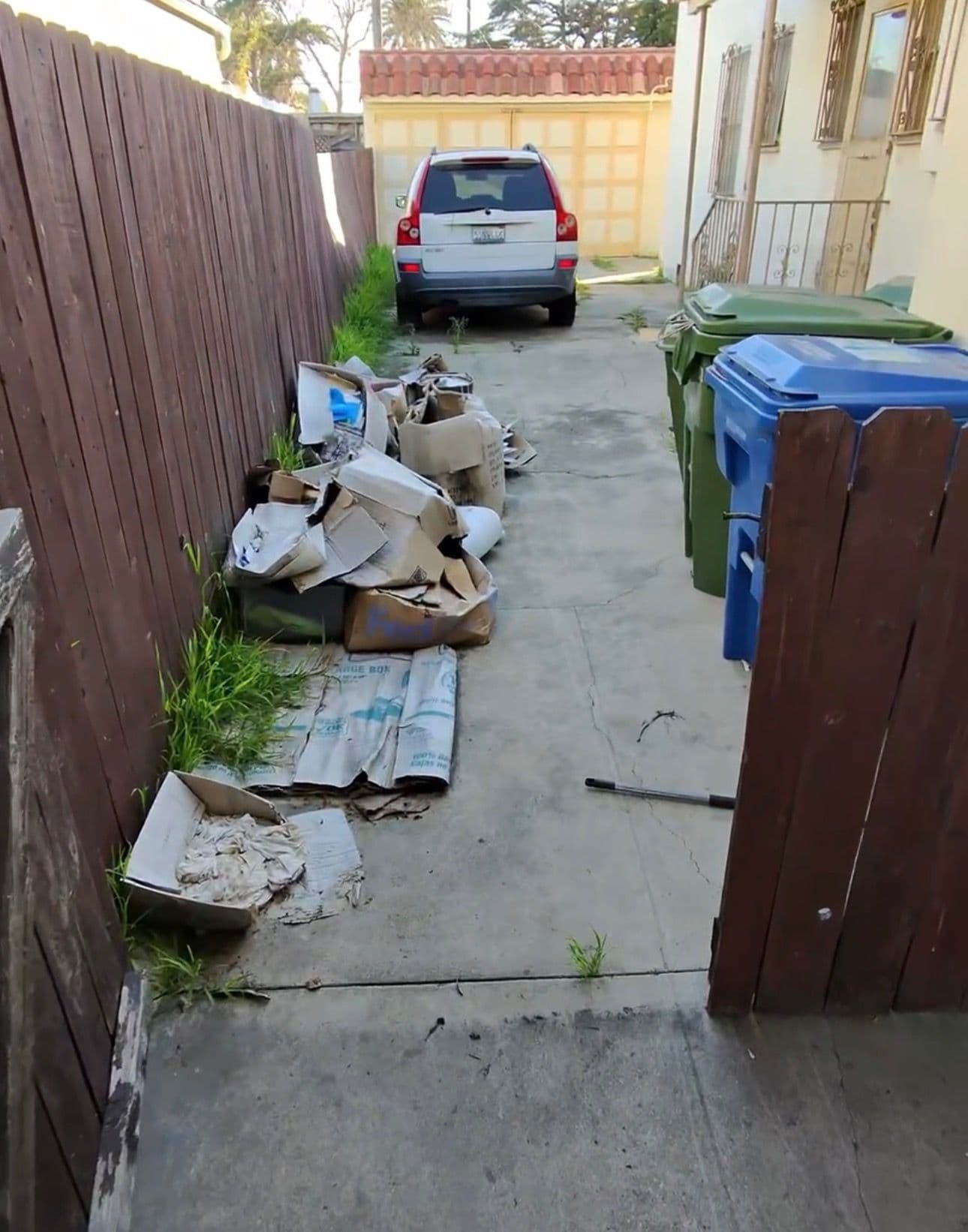 cluttered alleyway with cardboard waste and green recycling bins beside a parked vehicle