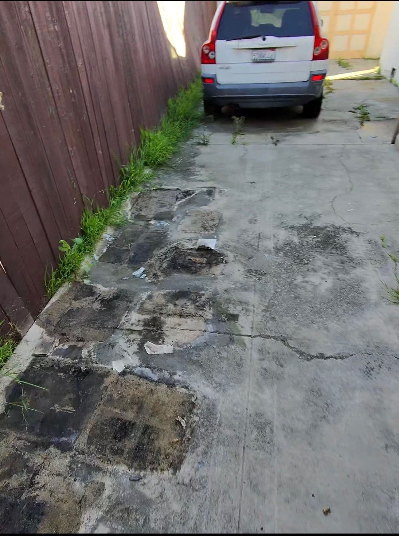 damaged driveway with overgrown grass and a parked car alongside a wooden fence